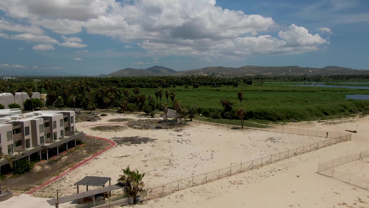 At 0:01 in San Jose del Cabo, the white resort building remains in the left foreground while fenced sand, scattered palms, green fields, distant hills, and no visible people fill the frame.