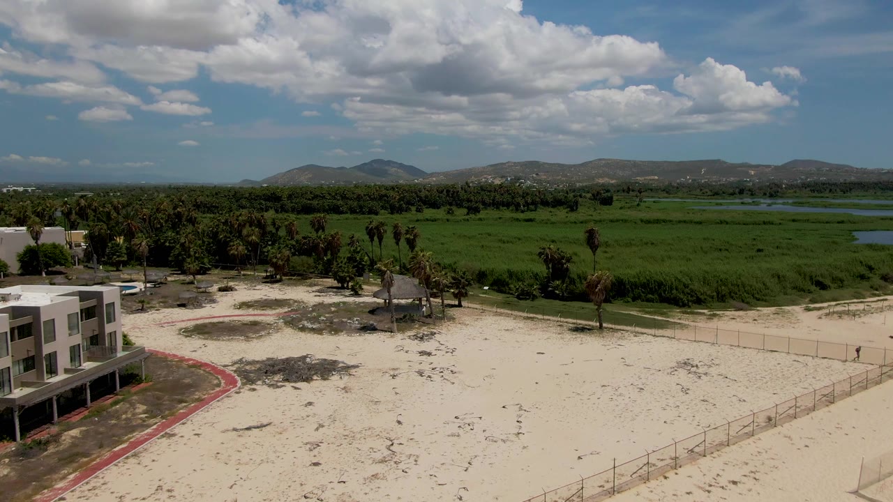 At 0:02 in San Jose del Cabo, the resort building has shifted farther left as the sandy fenced foreground, palms, green field, distant hills, blue sky, clouds, and no visible people dominate the view.