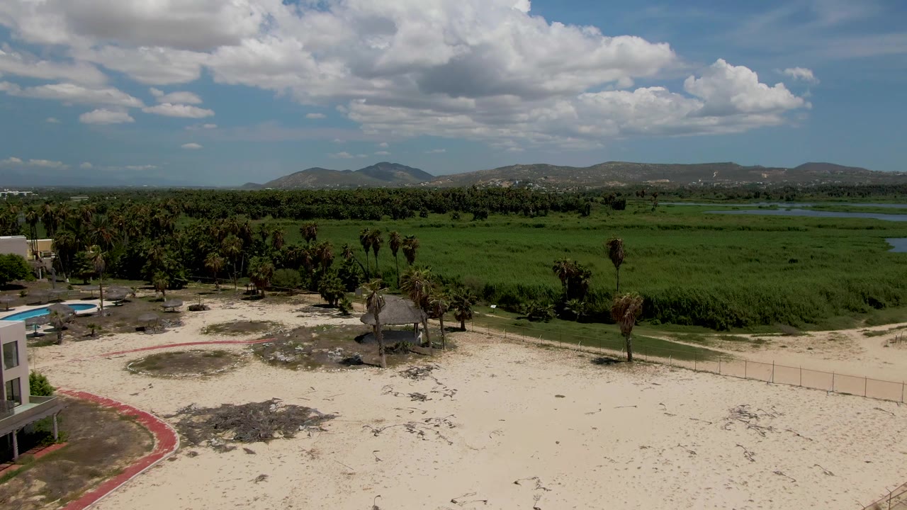 Partial resort building at left with sandy beach, palms, green field, hills, and clouds.