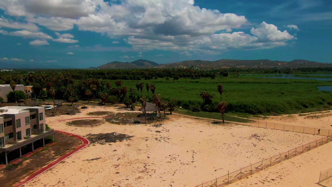 Aerial frame in San Jose del Cabo showing a partial modern building at left, sandy ground, palm trees, green fields, distant hills, blue water patches, and no visible people.