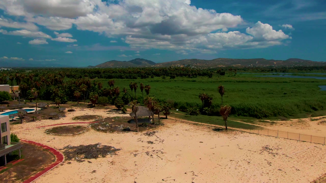 Aerial frame in San Jose del Cabo with the building nearly out of view, pale sand and palms in front, a fence line, green fields, distant hills, and no visible people.