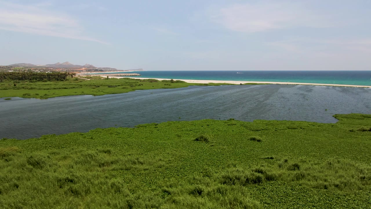 Mid-opening frame from above San Jose del Cabo, showing a broad green wetland edge, dark estuary water, pale sandbar, deep blue ocean, low hills, and no visible people.