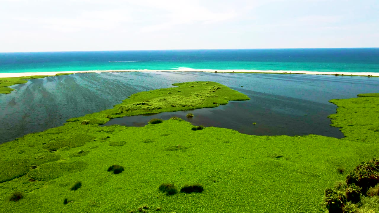 Aerial frame from San Jose del Cabo with the bright marsh foreground stretching across the lower frame, lagoon water on both sides, ocean surf behind the sandbar, and no visible people.