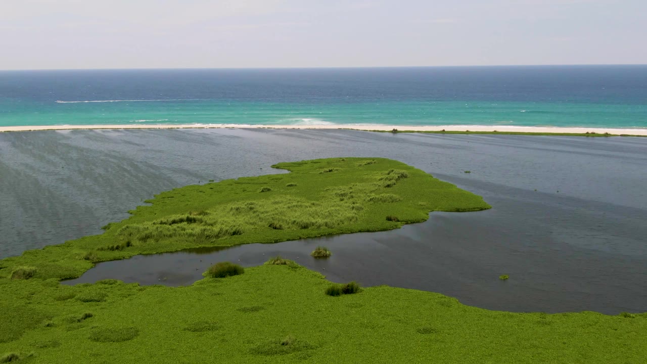 Aerial San Jose del Cabo coastal frame showing green marshland, dark lagoon water, a pale sandbar, turquoise ocean, faint surf, a small distant boat wake, and no visible people.