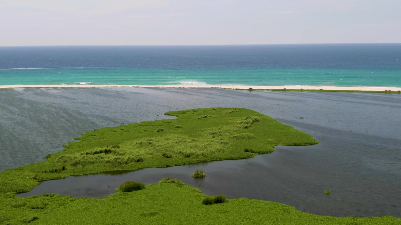 Aerial frame from San Jose Del Cabo showing a green island surrounded by dark lagoon water, with a white beach and turquoise ocean beyond, no visible people.