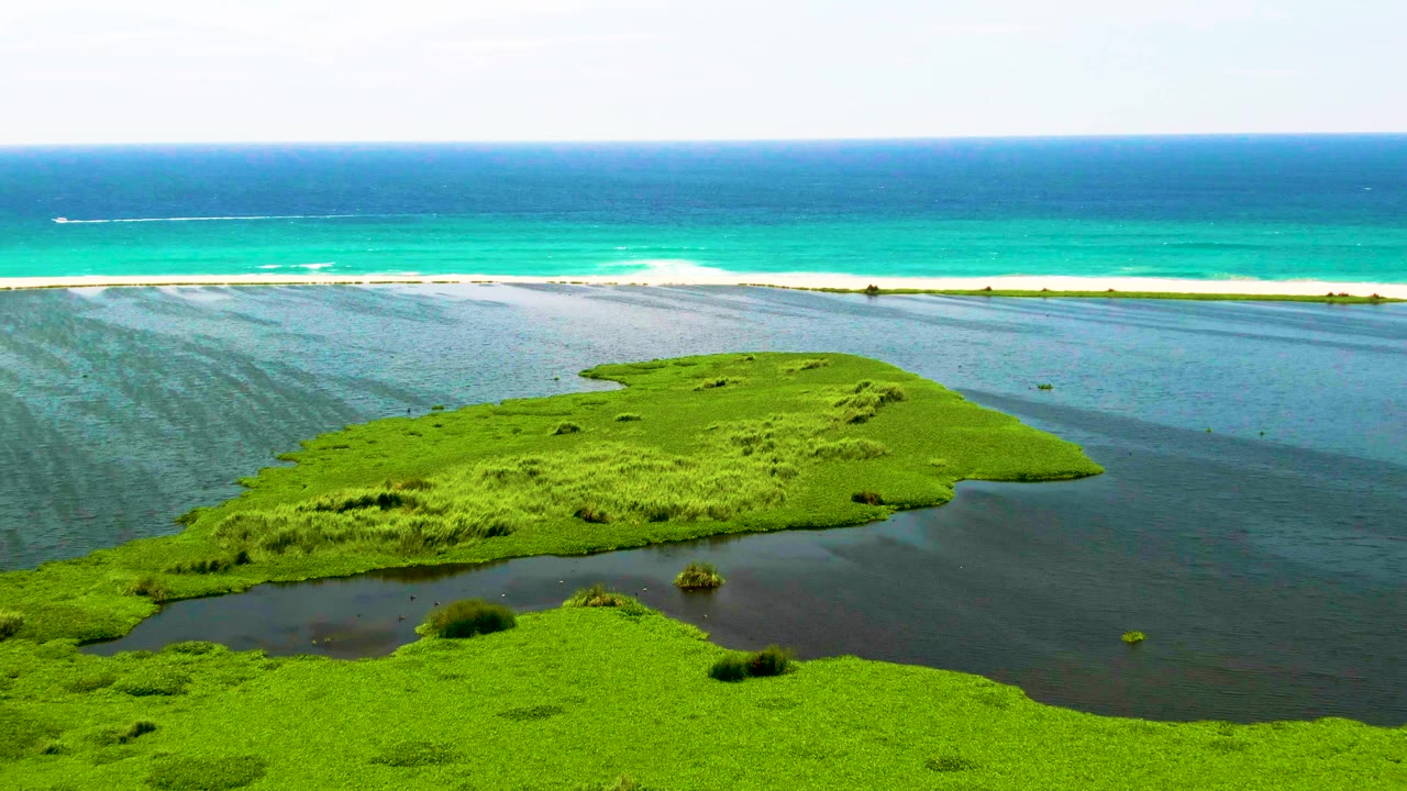 Aerial frame showing the central green lagoon island, sheltered dark water, white surf, and a small boat farther out on the ocean near San Jose del Cabo, Mexico, with no visible people.