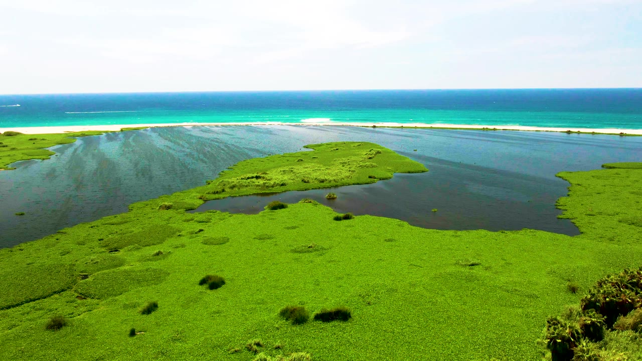 Aerial view in San Jose del Cabo with the central green landform extending into dark lagoon water, turquoise ocean bands beyond the beach, and no visible people.