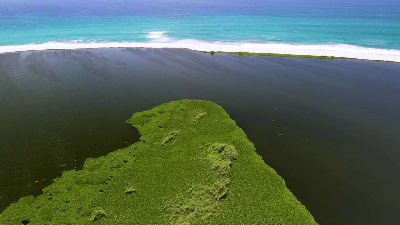 Mid-clip aerial view in San Jose del Cabo, Mexico, where the green landmass sits lower in frame beneath a broad dark water area, bright surf, blue-green ocean, and no visible people.