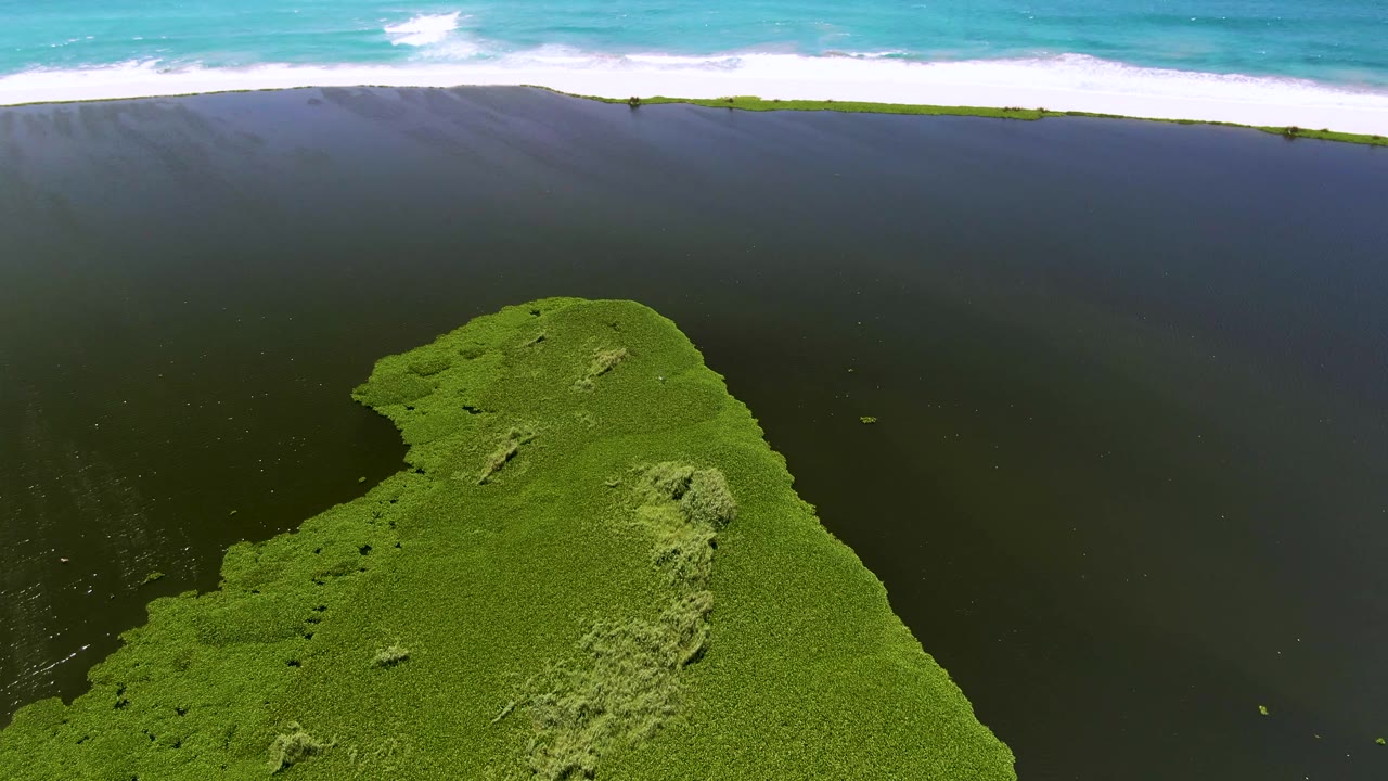 Aerial frame from San Jose Del Cabo, Mexico, with the green landmass shifted slightly lower, dark open water in midframe, white waves across the top, and no visible people.