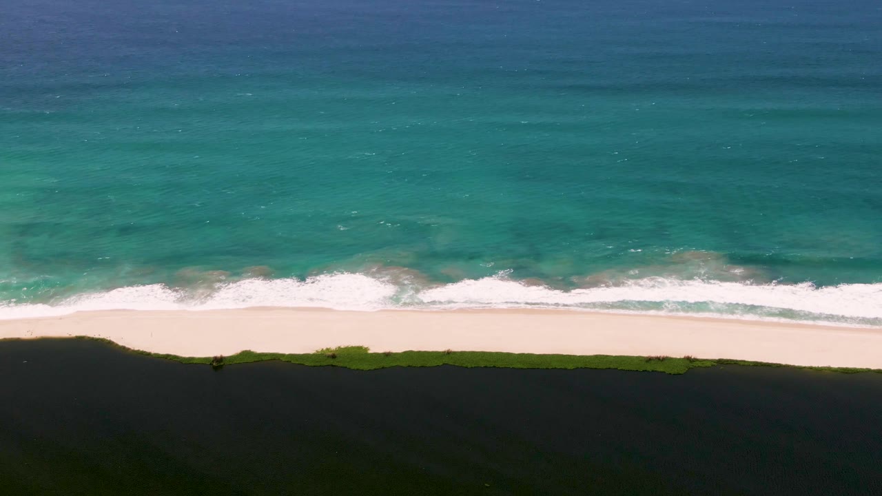 White waves break along a pale beach beside turquoise water and dark shadow