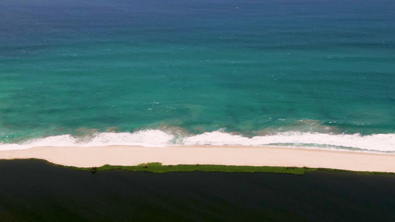 Overhead San Jose del Cabo beach frame with white foam reaching the sand, turquoise ocean above, green vegetation and dark shadow below, and a tiny person walking near the shoreline.