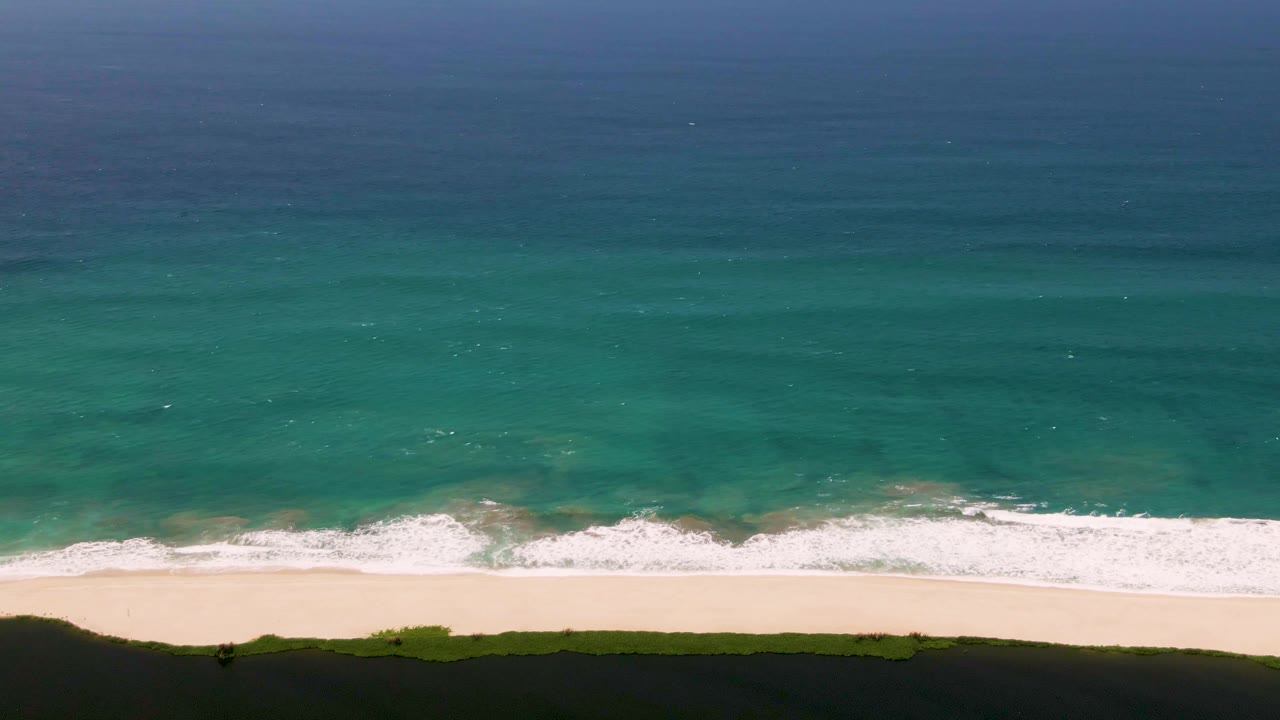 Aerial view over San Jose del Cabo shoreline with deep blue water fading to turquoise, white surf at the sand, a dark green coastal strip, and no visible people.