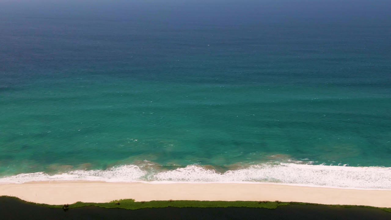 San Jose del Cabo surf line seen from above, with a brighter band of foam spreading across pale sand and open blue-green water filling most of the frame, no visible people.