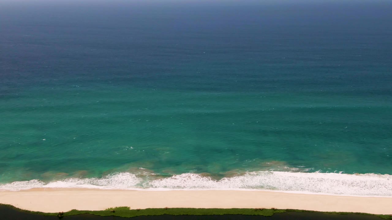 High aerial frame of the San Jose del Cabo coast with layered blue ocean, turquoise shallows, a thin white surf band, pale beach, and no visible people or structures.
