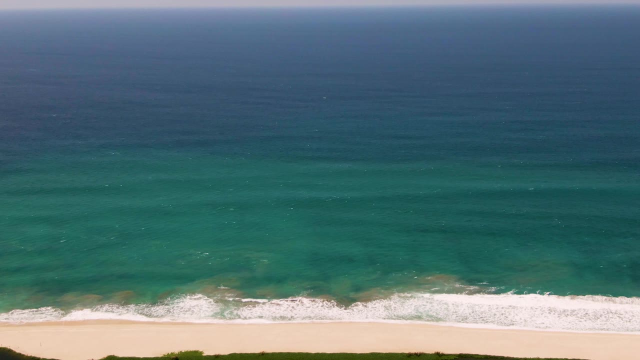Aerial San Jose del Cabo shoreline frame where the surf sits lower in the image, with broad teal water above, pale sand below, and no visible people.