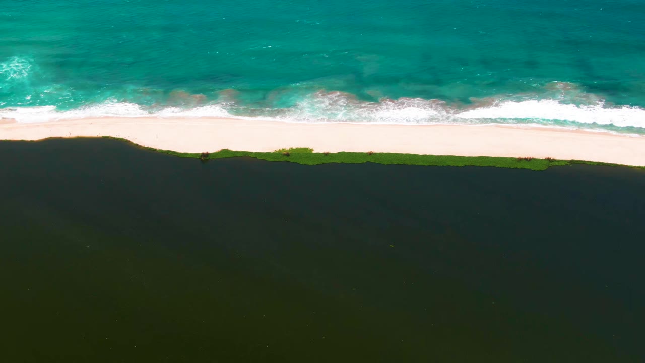 At 0:00, an overhead view in San Jose del Cabo shows turquoise ocean, a thin white surf line, pale beach sand, green vegetation, and darker dunes with no visible people.