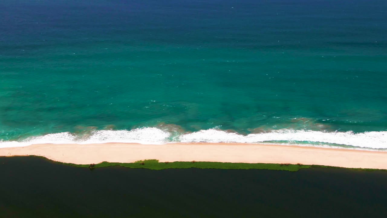 Overhead view of San Jose del Cabo shoreline with deep blue ocean, turquoise shallows, white surf, pale sand, dark dunes, and no visible people.