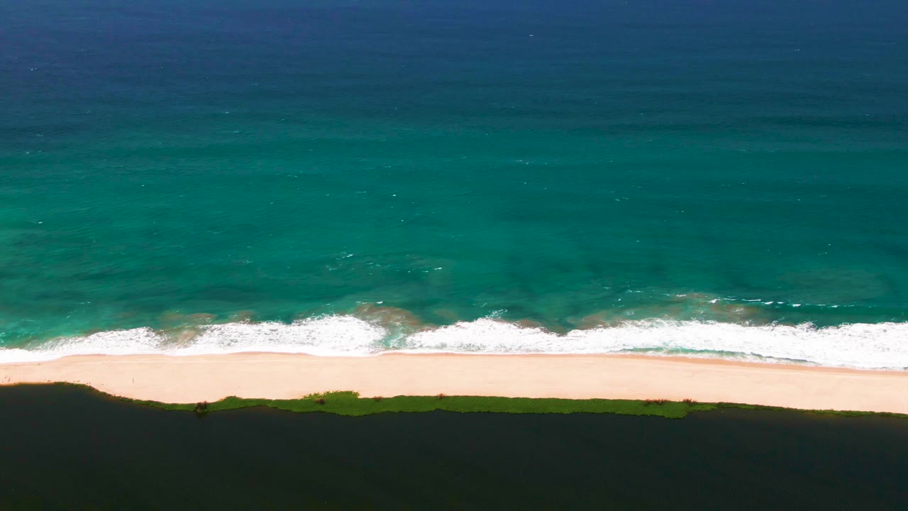 Aerial frame over San Jose del Cabo beach where a wide band of white foam rolls toward pale sand beneath blue-green water, with no visible people.