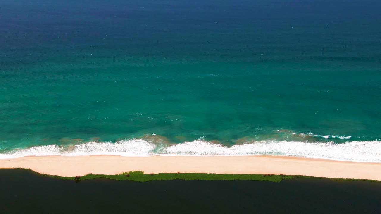 Top-down San Jose del Cabo coastline frame with turquoise water above a foamy surf band, light sand in the middle, dark dunes below, and no visible people.