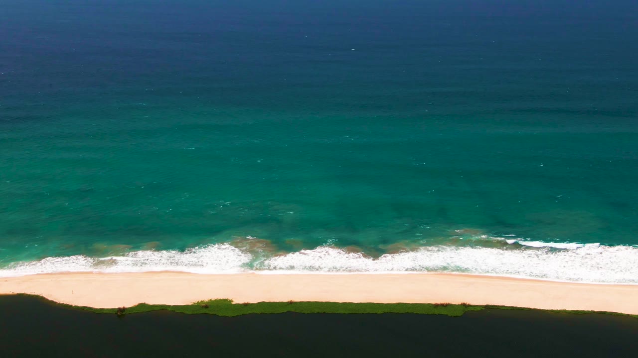 San Jose del Cabo aerial shoreline with blue water fading into green shallows, fresh white wave foam crossing the beach edge, dark dunes, and no visible people.