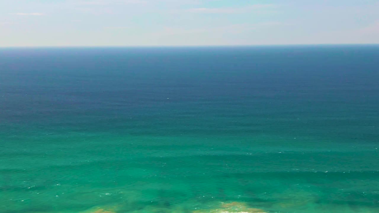 Aerial view in San Jose Del Cabo, Mexico, with blue-green ocean under a pale sky and a thin line of white surf near the bottom, no visible people.