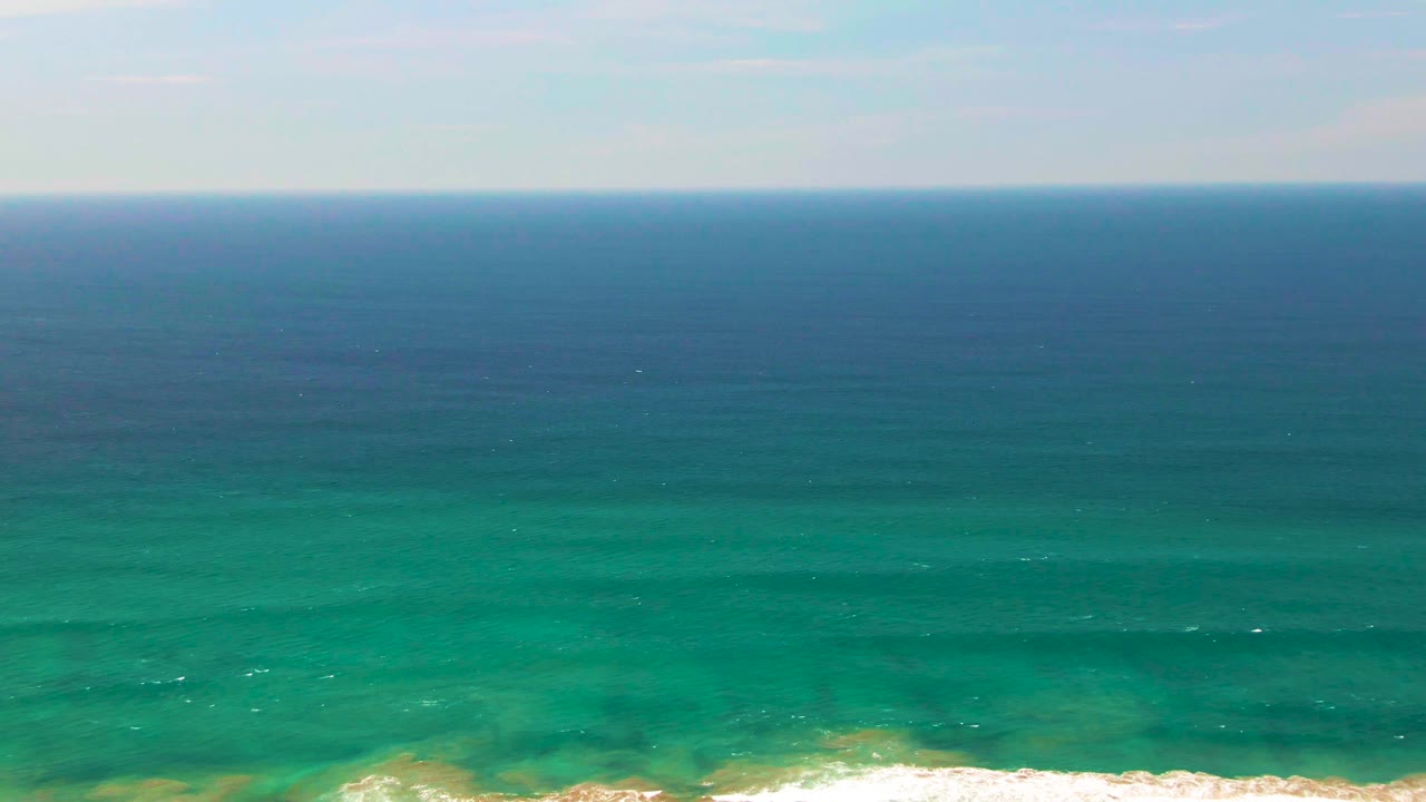 Aerial coastal frame from San Jose Del Cabo showing turquoise water, darker blue sea near the horizon, and white waves forming along the lower edge, no visible people.
