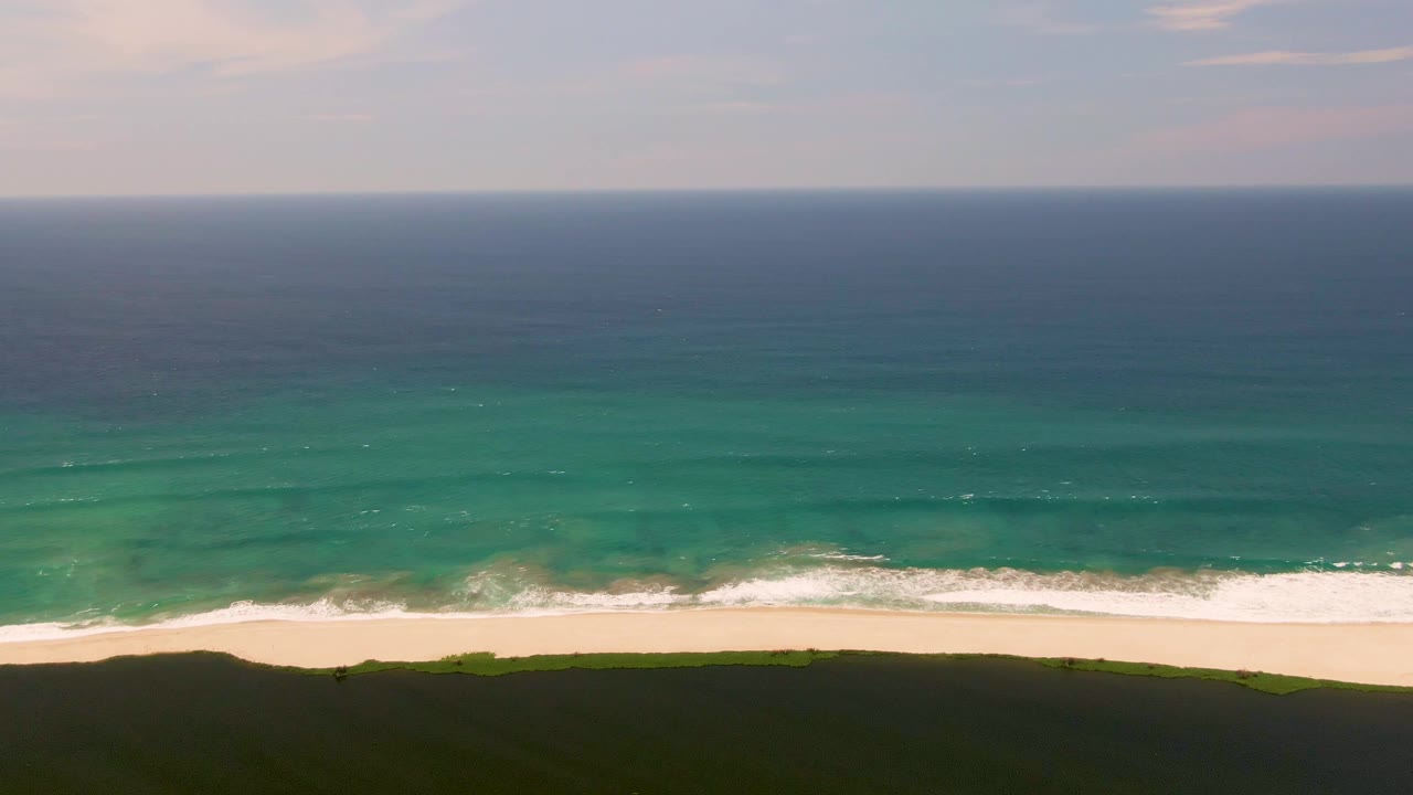 Aerial view of turquoise ocean waves reaching a sandy beach beside a dark grassy dune.