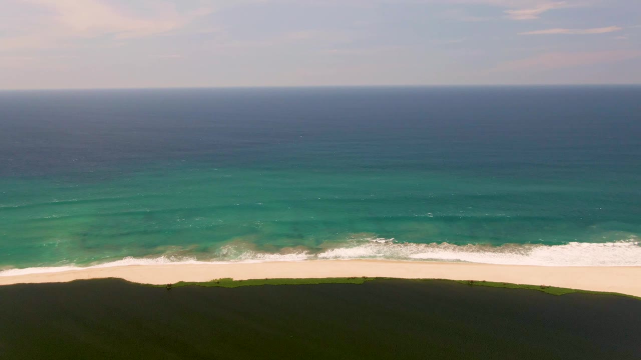 The San Jose del Cabo coast shows a slightly shifted white wave line breaking along the sand, with turquoise water offshore, a dark green dune below, and no visible people.