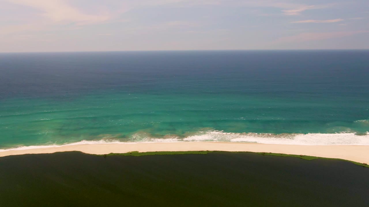 A wide aerial frame of San Jose del Cabo shoreline places the dark grassy dune across the lower edge, the pale sand and broken foam in the center, and blue ocean under a lightly clouded sky, with no visible people.