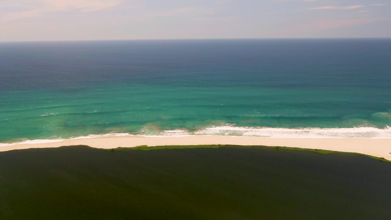 At the opening moment in San Jose del Cabo, turquoise ocean bands approach a white sand beach bordered by a dark green dune, with no visible people.