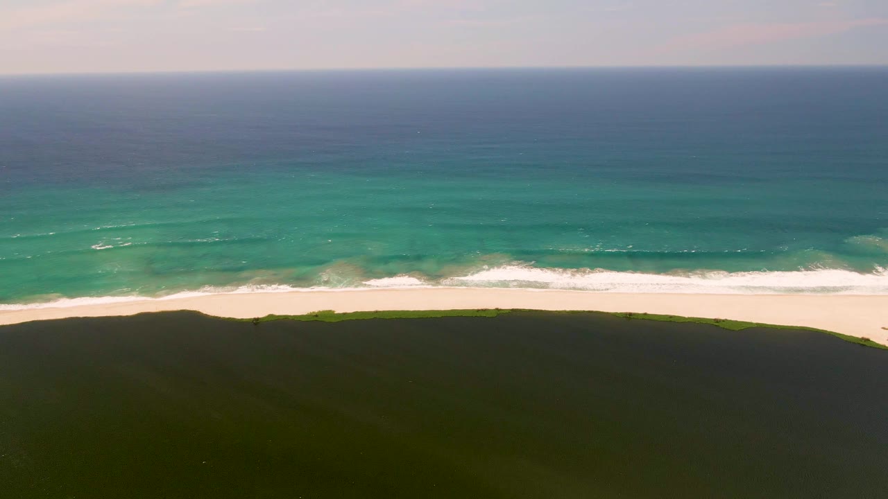 Aerial shoreline with green dune foreground, white sand, and layered blue ocean.