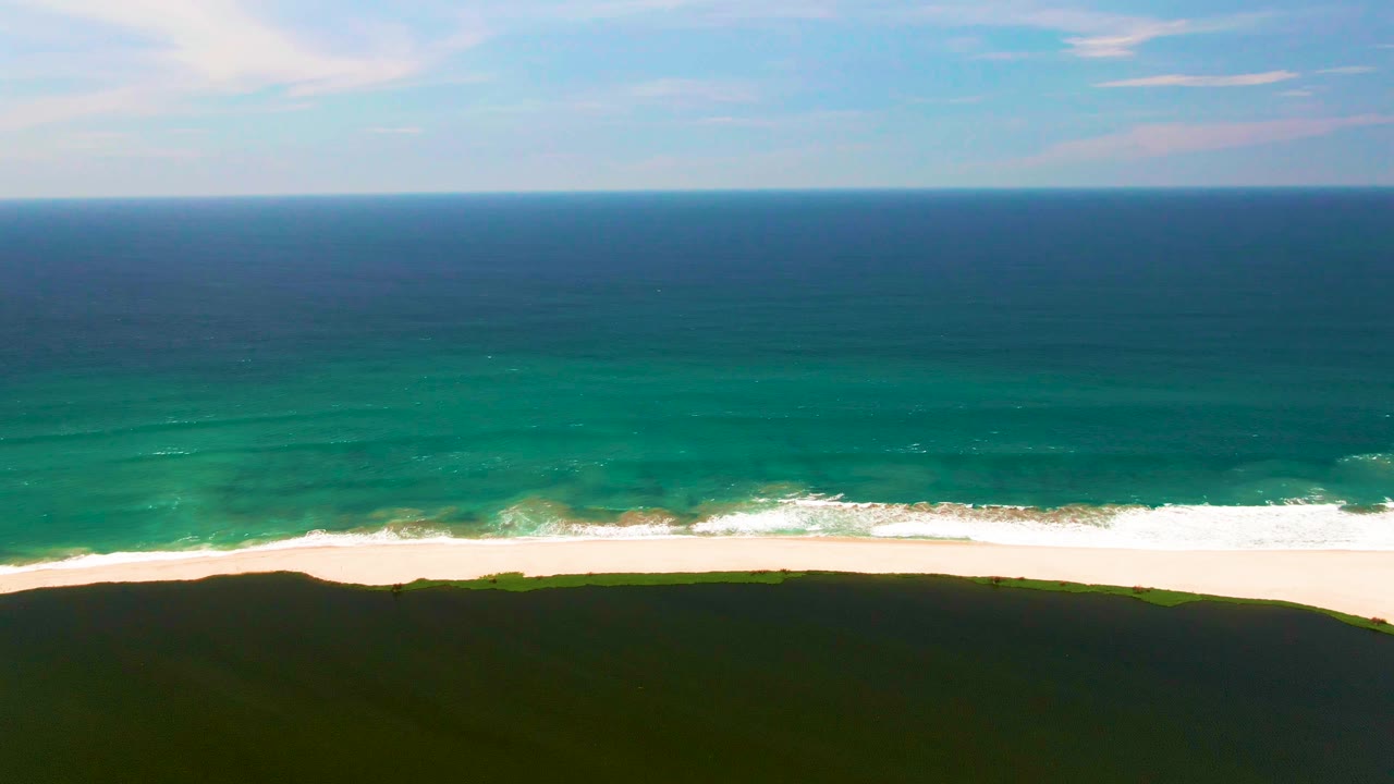 Aerial frame of San Jose del Cabo coastline with deep blue ocean, turquoise shore water, a bright surf line, dark green dunes in the foreground, and no visible people.