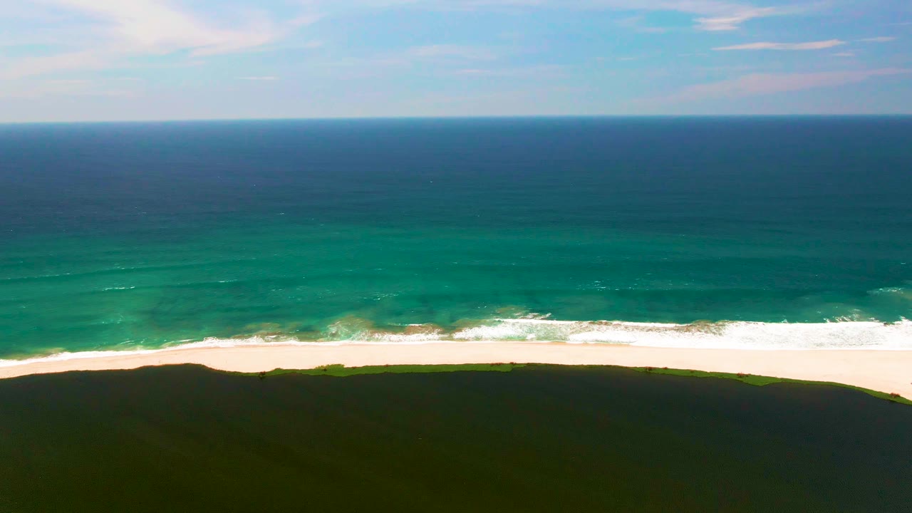San Jose del Cabo beach seen from above as the turquoise band nears the sand and white foam spreads along the shoreline, with green dunes below and no visible people.