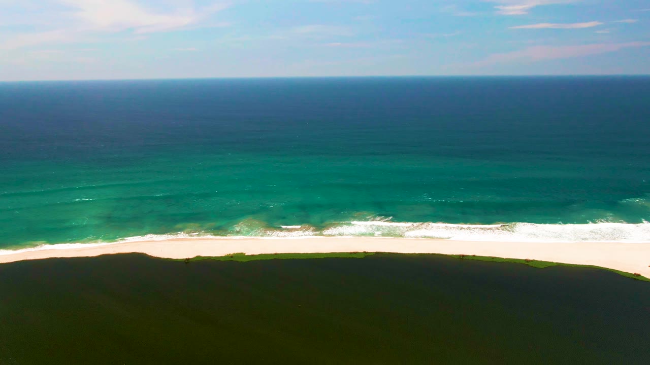 Aerial view of the San Jose del Cabo shoreline with a level ocean horizon, darker offshore water, lighter turquoise shallows, a white wave band, and no visible people.