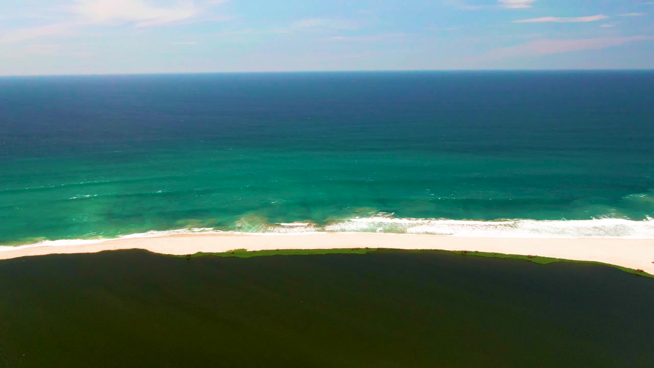 Aerial beach frame with white foam moving across sand and dark green dune foreground.