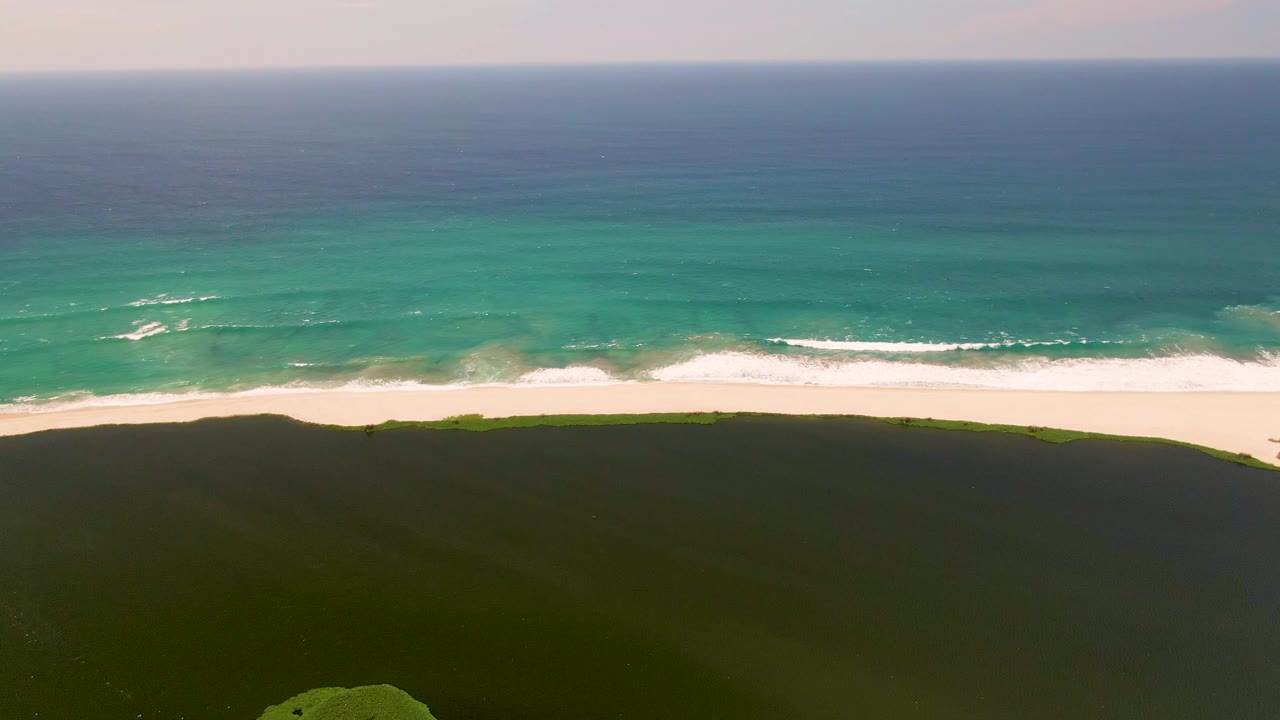 Second aerial frame of the San Jose del Cabo coast, with the white surf band slightly shifted along the beach, green dunes filling the lower frame, turquoise water offshore, and no visible people.