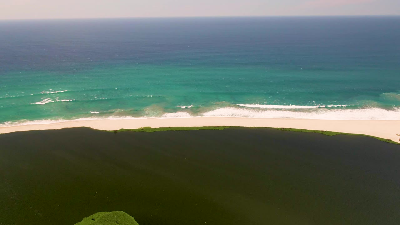 Third aerial frame over San Jose del Cabo coastal dunes, where the dark foreground contrasts with a bright sand strip, patchy whitecaps, turquoise shallows, blue ocean, and no visible people.