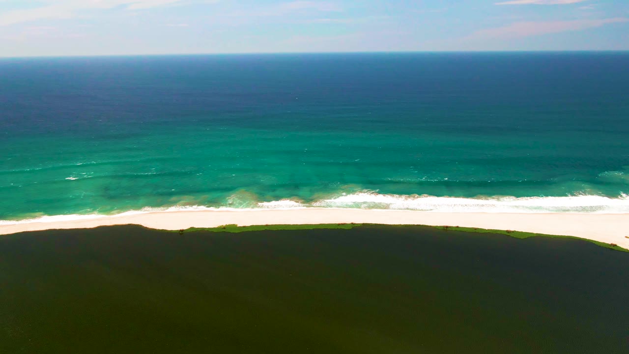 Opening frame of San Jose del Cabo coastline with turquoise water meeting a white sandy shore, dark green dunes below, a level horizon above, and no visible people.
