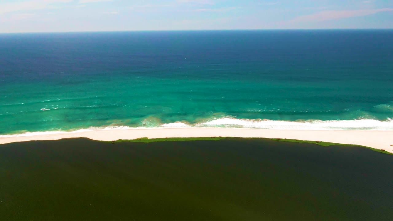 San Jose del Cabo aerial shoreline view with small white wave lines shifting near the center, bright sand across the frame, dark green dunes below, and no visible people.