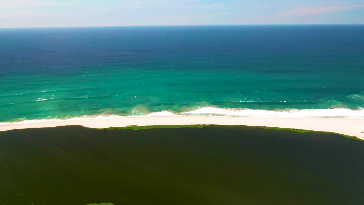 San Jose del Cabo coast seen from above with darker blue water near the horizon, teal water near shore, a continuous white beach, dark dunes, and no visible people.