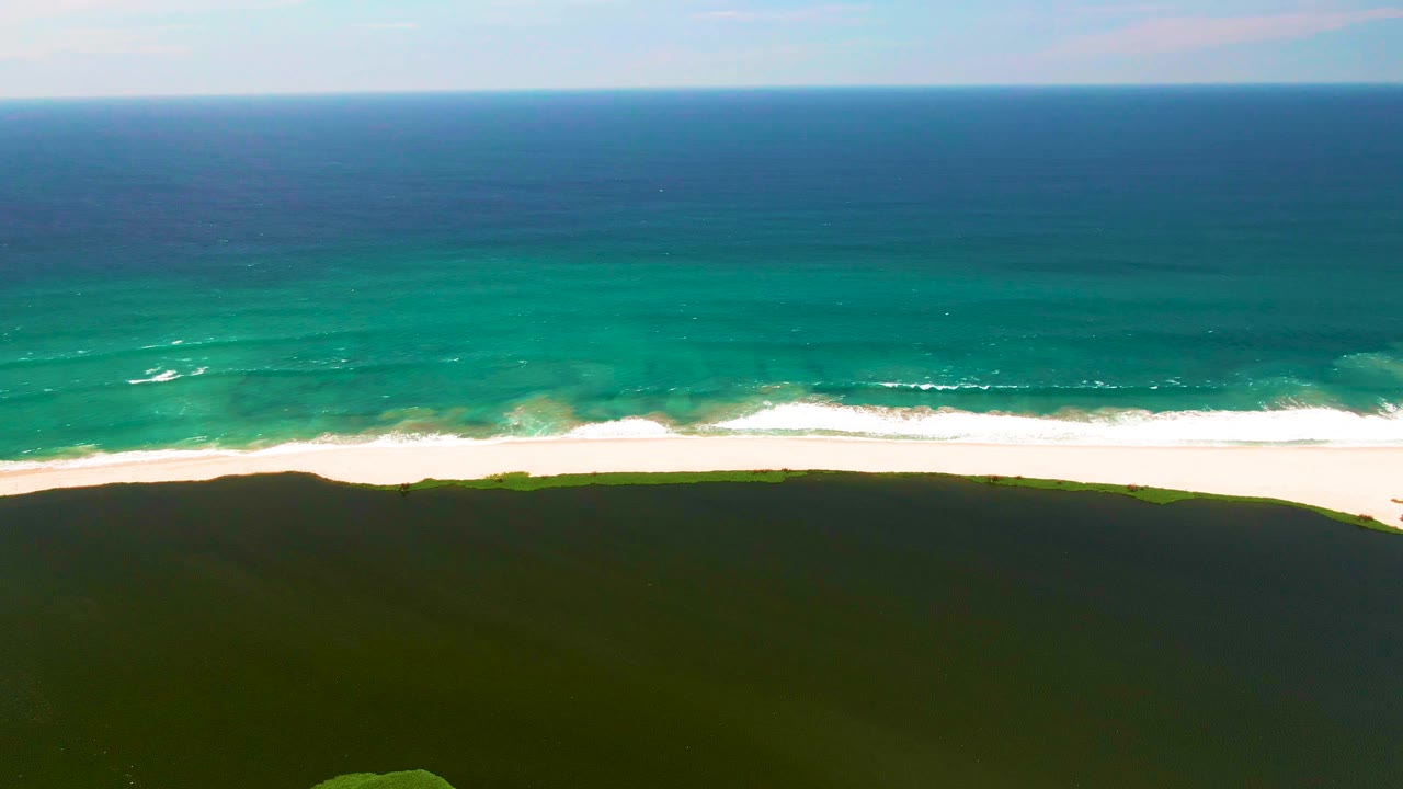 San Jose del Cabo shoreline frame with a low, even horizon, turquoise surf advancing toward the bright beach, dark green dune cover in the foreground, and no visible people.