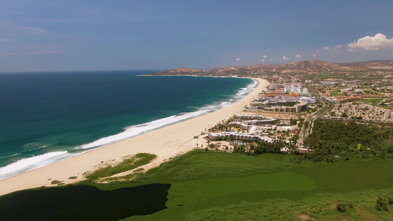 Opening aerial view over San Jose Del Cabo coastline with blue ocean, white surf, sandy beach, coastal buildings, green foreground, distant hills, and no visible people.