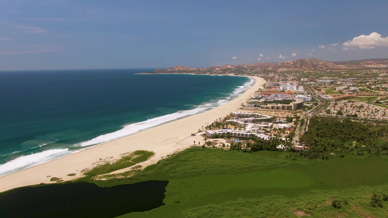 One second into the San Jose Del Cabo aerial, the surf line is slightly farther along the beach with coastal buildings and green vegetation visible, no visible people.