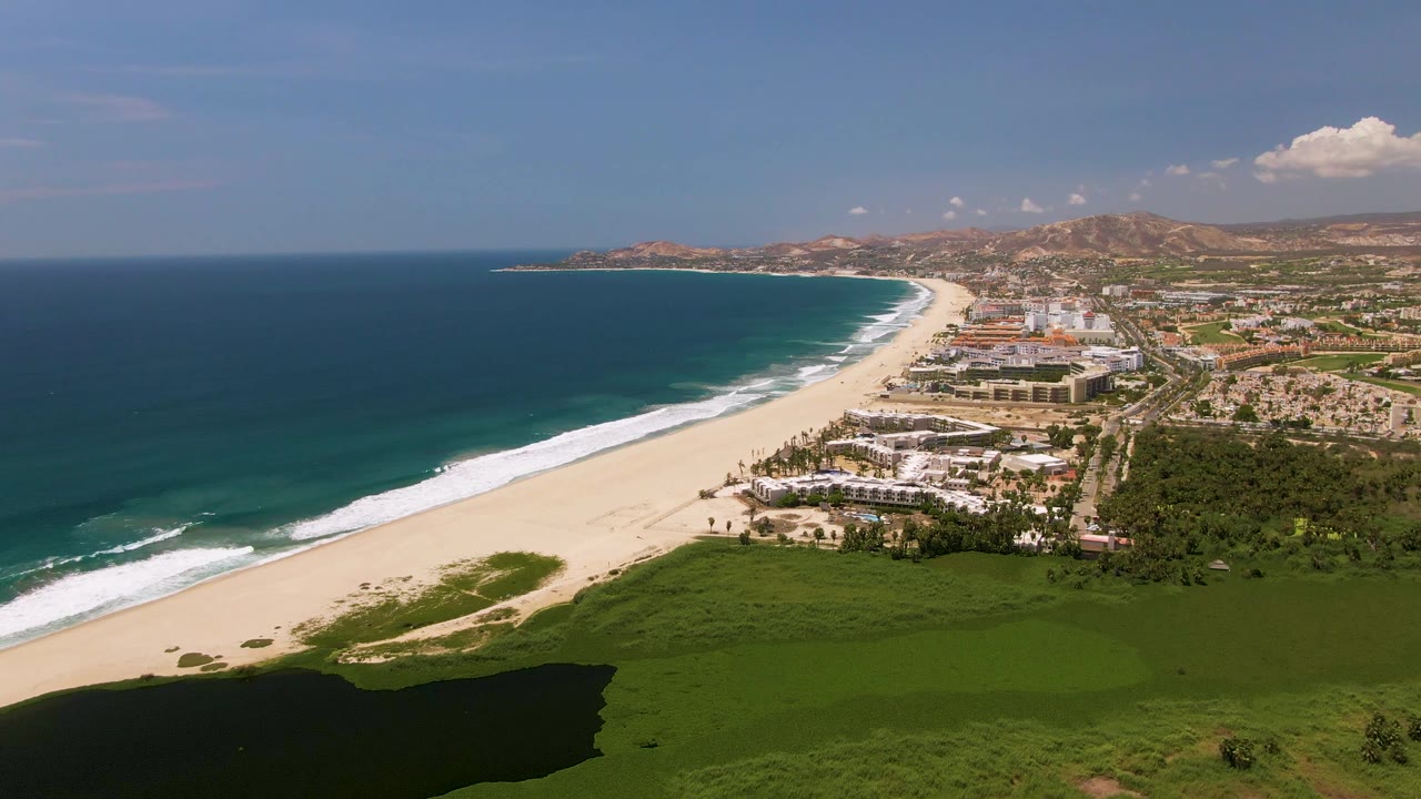 At 0:02, the San Jose Del Cabo beach remains centered between dark ocean and coastal buildings, with a broad green foreground and no visible people.