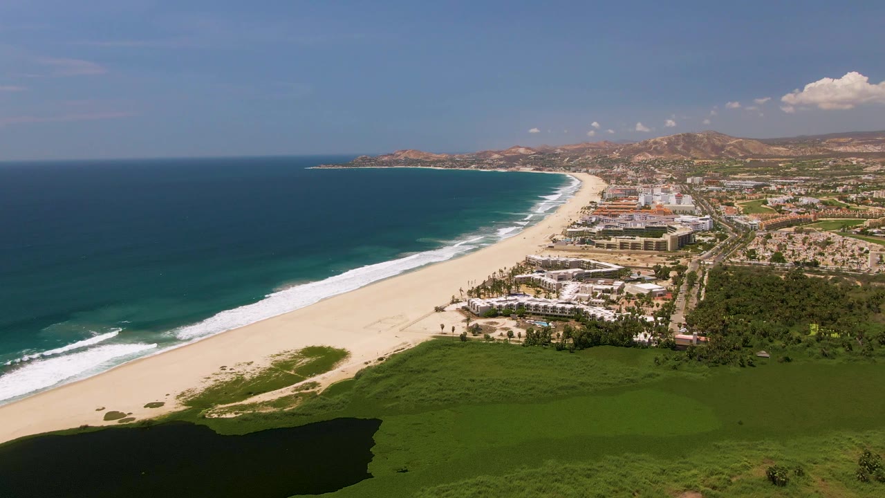 Aerial beach scene with ocean waves, pale sand, resort-like buildings, greenery, and a shadowed foreground.