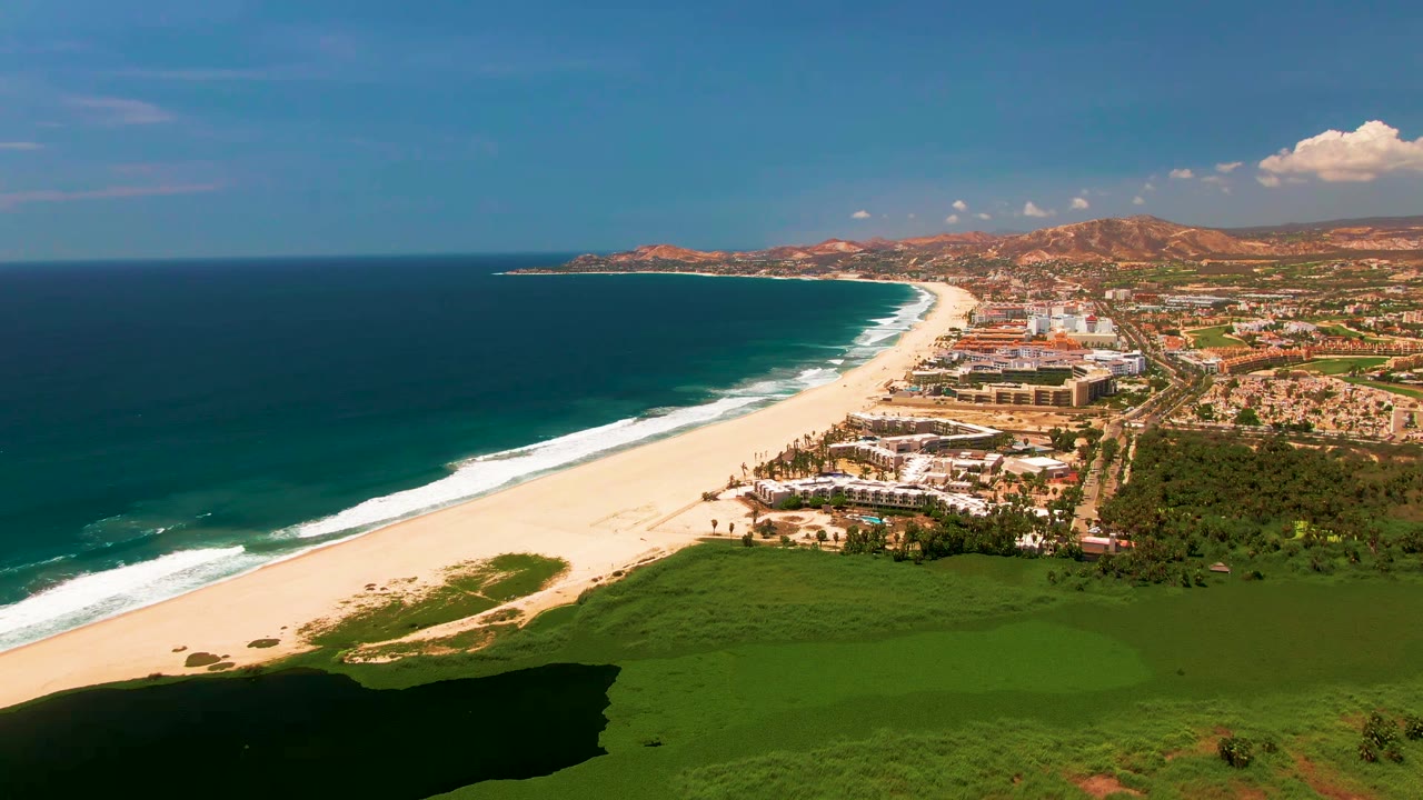 Opening aerial frame over San Jose Del Cabo with a dark shadow crossing the green field, white beach and surf to the left, beachfront buildings in the center, distant hills, and no visible people.