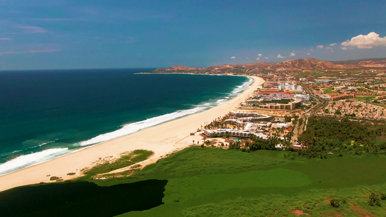 At 0:01, the San Jose Del Cabo aerial view holds on the same beach curve as waves advance along the shore beside the green field and beachfront buildings, with no visible people.