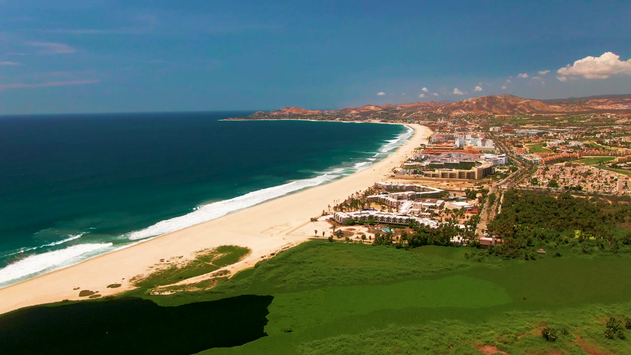 At 0:02, the aerial frame shows turquoise water, a long white beach, clustered beachfront buildings, green foreground land, reddish hills in the distance, and no visible people.