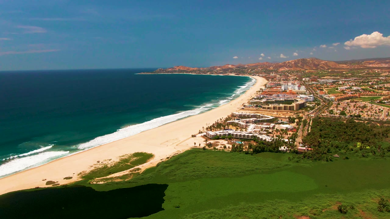 Aerial coastline with surf, sandy beach, buildings, and shadowed green field.