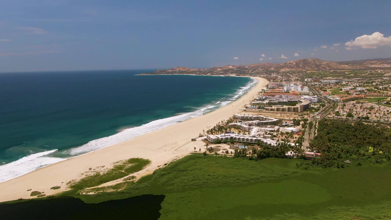Aerial view over San Jose Del Cabo beach with blue ocean on the left, a broad sandy shoreline, green foreground vegetation, coastal buildings, inland hills, and no visible people.